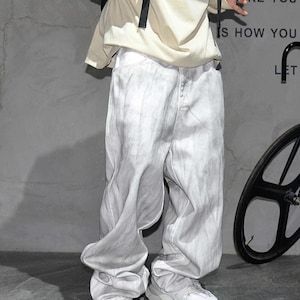 May include: Light gray denim pants with a unique, flowing design. The pants have a faded, almost tie-dye effect, and are paired with white sneakers. The overall style is casual and modern.