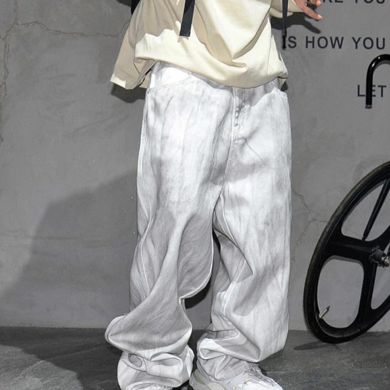 May include: Light gray denim pants with a unique, flowing design. The pants have a faded, almost tie-dye effect, and are paired with white sneakers. The overall style is casual and modern.