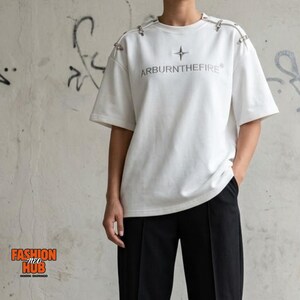 May include: A white short-sleeve t-shirt featuring a silver star graphic and the text ARBURNTHEFIRE printed on the chest. The shoulders are accented with silver metal hardware details. The shirt is paired with black trousers.