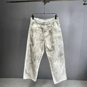 May include: Light gray, wide-leg jeans with a tie-dye effect hanging on a black hanger. The jeans have a classic five-pocket design and a button and zipper closure. The fabric appears to be a medium-weight denim.