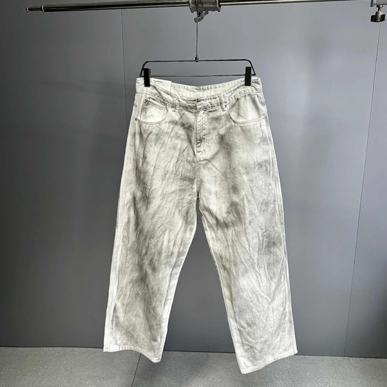 May include: Light gray, wide-leg jeans with a tie-dye effect hanging on a black hanger. The jeans have a classic five-pocket design and a button and zipper closure. The fabric appears to be a medium-weight denim.