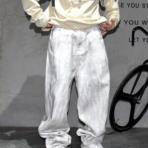 May include: Light gray, baggy-fit jeans with a faded, textured appearance. The jeans have a relaxed, oversized silhouette. The model is wearing a cream-colored long-sleeve shirt. The background is a plain gray wall.