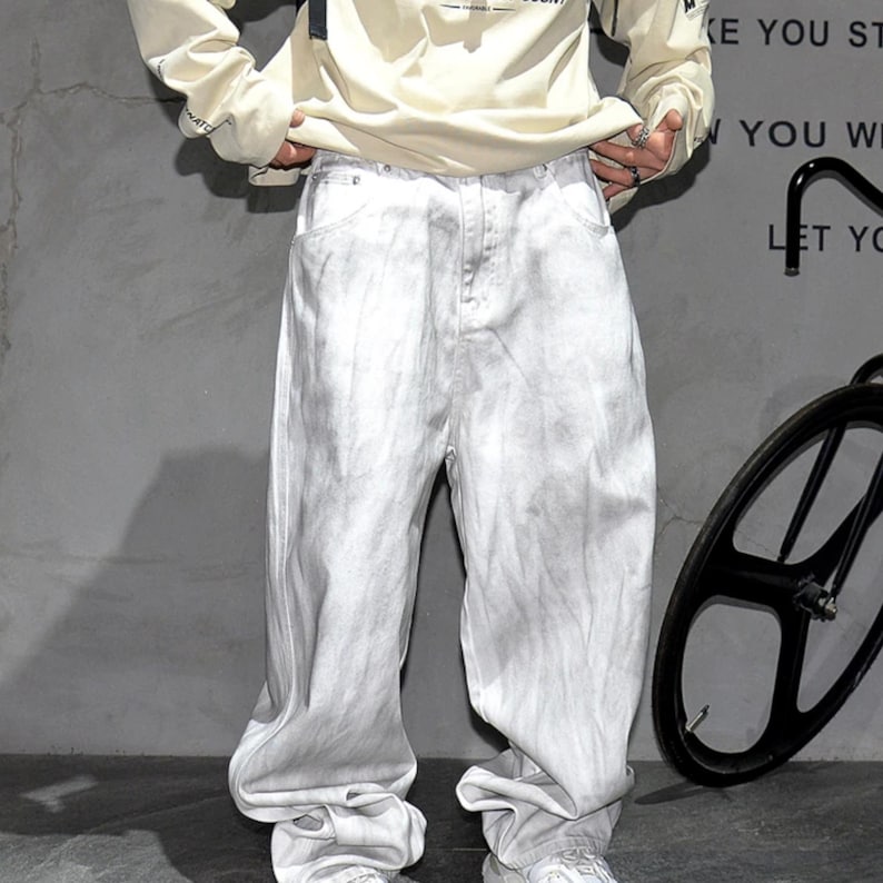 May include: Light gray, baggy-fit jeans with a faded, textured appearance. The jeans have a relaxed, oversized silhouette. The model is wearing a cream-colored long-sleeve shirt. The background is a plain gray wall.
