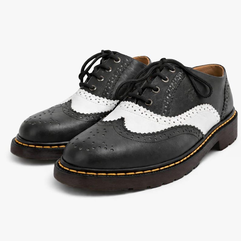 May include: Black and white leather oxford shoes with a wingtip design. The shoes have a lace-up closure, a thick brown sole, and yellow stitching around the sole. The shoes are a classic style.