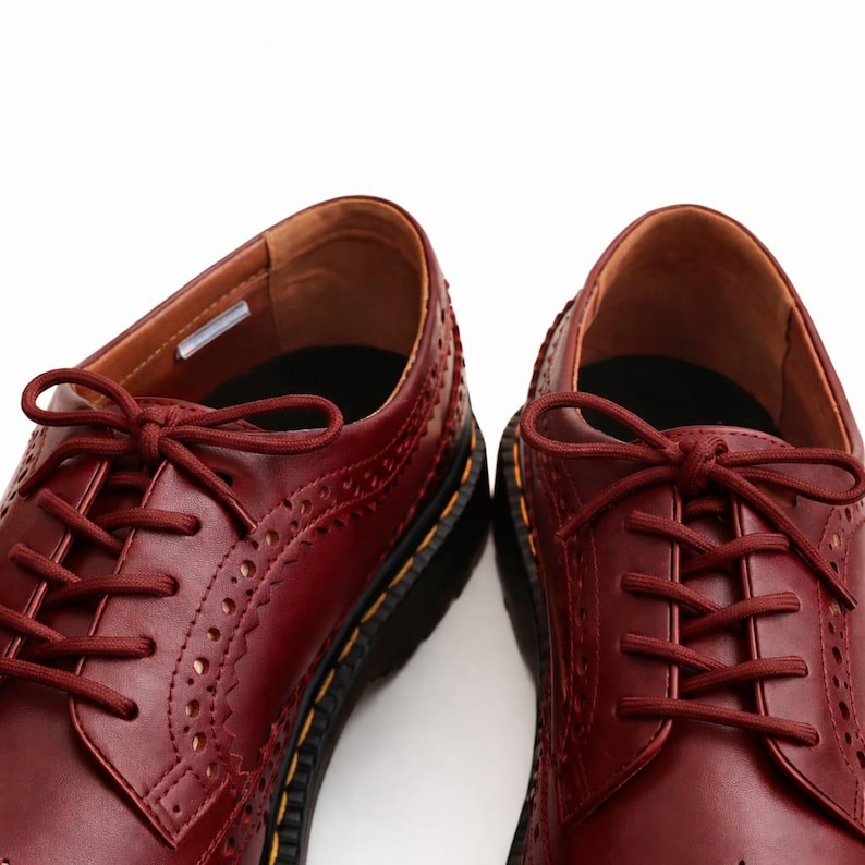 May include: A pair of burgundy leather Oxford shoes with decorative perforations and matching laces. The shoes have a classic design with a rounded toe and a thick black sole with yellow stitching. The interior is a lighter shade of brown.