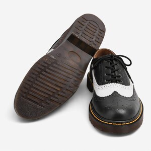 May include: Black and white leather wingtip shoes with a brown sole. The shoes have a classic design with a perforated pattern and a lace-up closure. The sole is thick and sturdy, with a visible yellow stitch.