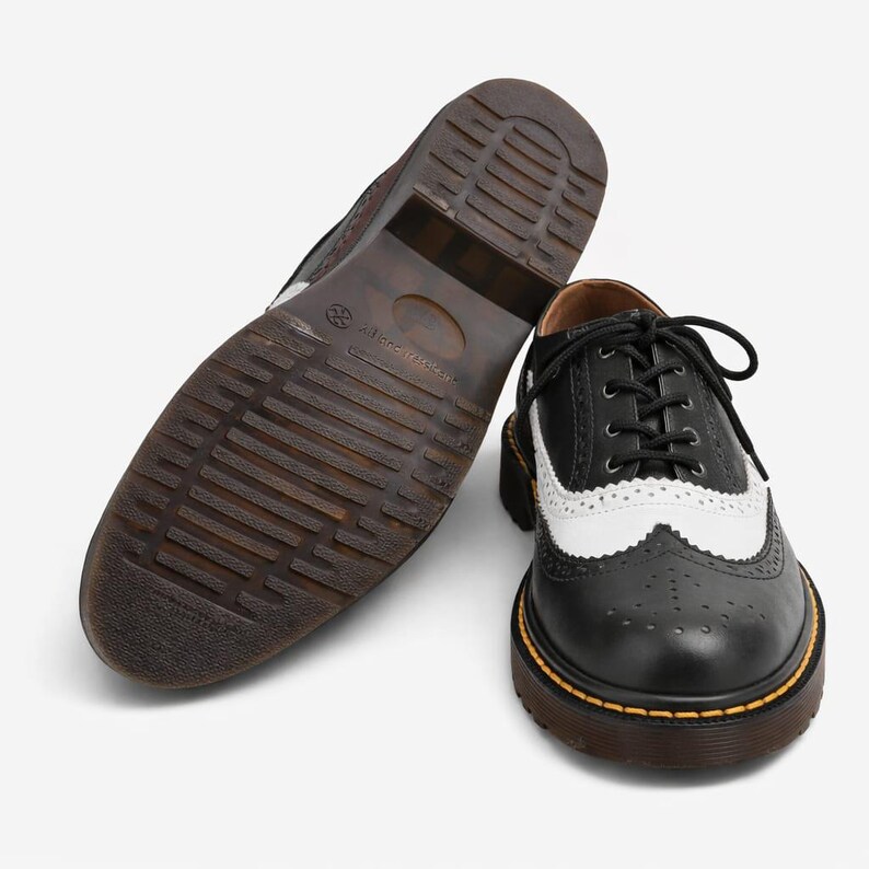 May include: Black and white leather wingtip shoes with a brown sole. The shoes have a classic design with a perforated pattern and a lace-up closure. The sole is thick and sturdy, with a visible yellow stitch.