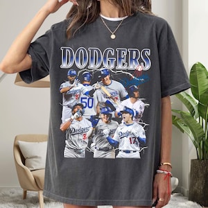 Può includere: Una t-shirt grigio antracite a maniche corte con una stampa grafica di diversi giocatori di baseball in divisa bianca. La parola DODGERS è stampata in grandi lettere blu e bianche nella parte superiore, con fulmini che circondano i giocatori.