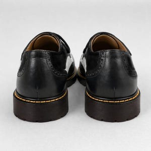 May include: Pair of black leather shoes with white accents and a thick, dark brown sole. The shoes have a classic design with decorative perforations and yellow stitching around the sole. The shoes are viewed from the back.