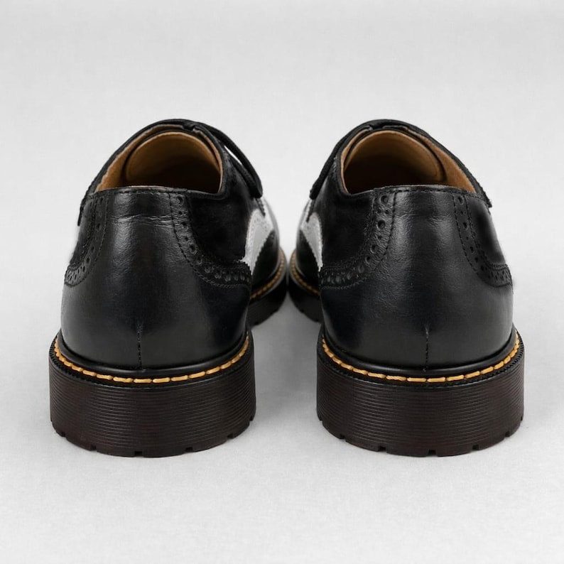 May include: Pair of black leather shoes with white accents and a thick, dark brown sole. The shoes have a classic design with decorative perforations and yellow stitching around the sole. The shoes are viewed from the back.