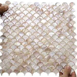 以下が含まれることがあります： 魚の鱗模様の正方形モザイクタイルシート。 タイルは真珠のような白で、ベージュとピンクの微妙な色合いがあります。 モザイクは両手で持たれており、タイルの形状とデザインを示しています。