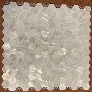 以下が含まれることがあります： 多数の虹色の六角形のタイルで構成された正方形のモザイクタイルシート。 タイルは主に白で、微妙な色の変化と質感があり、真珠のような効果を生み出しています。 タイルはハニカムパターンに配置されています。