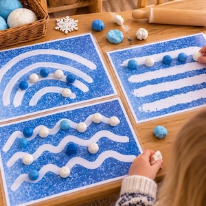 May include: Three laminated activity sheets with blue and white winter-themed patterns, including rainbows, horizontal lines, and wavy lines. Small blue and white modeling clay balls are placed along the printed paths, accompanied by wooden snowflake cutouts and a wooden rolling pin.