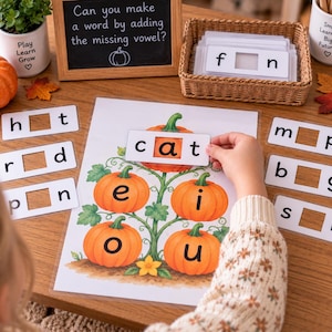 May include: An educational activity set featuring a laminated sheet with pumpkin illustrations and vowel letters, accompanied by word cards with missing vowels, a small chalkboard with the text Can you make a word by adding the missing vowel?, and a woven basket.