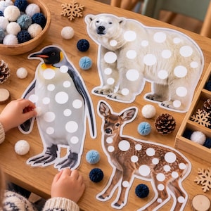 May include: Three animal cutouts of a polar bear, a penguin, and a deer featuring white circular spots, accompanied by a wooden bowl and a divided tray filled with small blue, white, and navy pom-poms, plus wooden snowflake and pinecone accents.