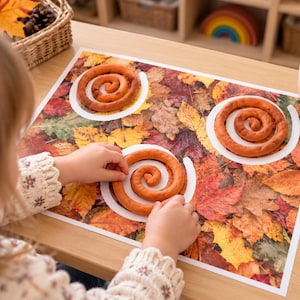 Op de afbeelding: Een rechthoekige speelmat met een hoge resolutie print van gevallen herfstbladeren in tinten rood, oranje, geel en bruin. De mat bevat drie witte cirkelvormige contouren die bedoeld zijn voor het plaatsen van oranje kleispiralen.