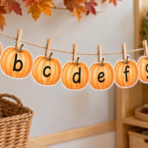May include: A decorative garland featuring eight orange pumpkin cutouts hanging from a brown twine string with wooden clothespins. Each pumpkin displays a single lowercase letter of the alphabet in black, starting with a and ending with h.