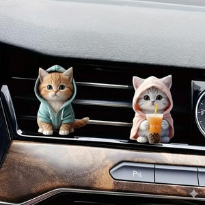 May include: Two cat figurines in hoodies. One is orange and white in a teal hoodie, the other is gray and white in a pink hoodie, holding a drink with the word "RAW" on the cup. The figurines are car air vent decorations.