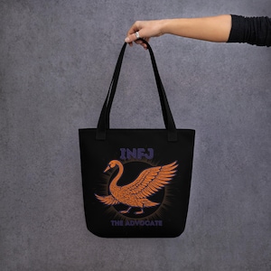 Könnte beinhalten: Schwarze Tragetasche mit einem orangefarbenen Schwan-Design, lila Umriss und dem Text "INFJ" oben und "THE ADVOCATE" unten. Der Schwan ist vor einem kreisförmigen Sonnenstrahl-Design platziert. Die Tasche hat zwei schwarze Griffe.
