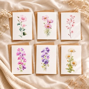 Könnte beinhalten: Sechs florale Grußkarten mit Aquarellillustrationen verschiedener Blumen, darunter rosa Kosmeen, Gänseblümchen und lila Blüten. Jede Karte liegt auf einem braunen Umschlag, angeordnet auf einer beigefarbenen Stoffoberfläche.