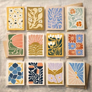 Może przedstawiać: Zestaw dwunastu kartek okolicznościowych z motywami kwiatowymi. Każda karta przedstawia unikalną ilustrację kwiatów i liści w odcieniach żółci, pomarańczy, błękitu i fioletu. Kartki ułożone są na neutralnej powierzchni, z brązowymi kopertami.