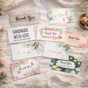 May include: A selection of thank you cards with floral and botanical designs. Some cards feature the text "Thank You" in elegant script, while others include phrases like "Handmade with Love". The cards are arranged on a white fabric surface.