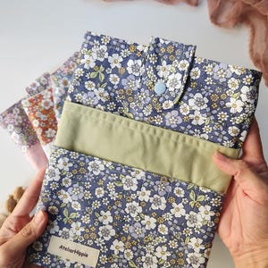 May include: A floral print fabric case with a light blue button closure. The case is navy blue with white and yellow flowers. The case has a light green band across the middle. The label says "AtelierHippie".