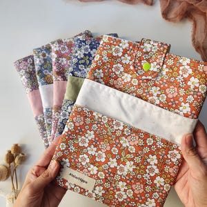 May include: A collection of floral fabric pouches in various colors, including orange, blue, and pink. Each pouch features a different floral pattern and a white accent. The pouches are held in a person's hands, with the label "AtelierHippie" visible.