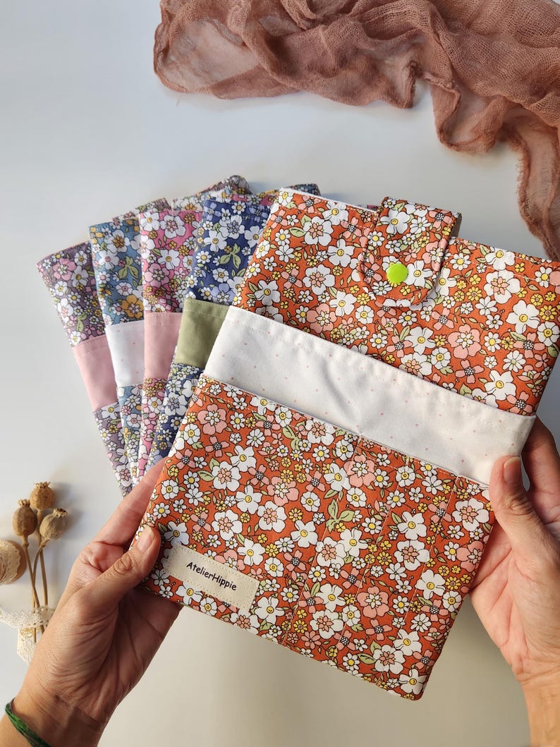 May include: A collection of floral fabric pouches in various colors, including orange, blue, and pink. Each pouch features a different floral pattern and a white accent. The pouches are held in a person's hands, with the label "AtelierHippie" visible.