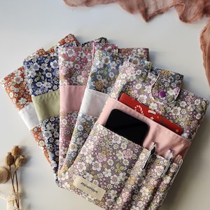 May include: A collection of floral fabric pouches in various colors, including orange, navy, pink, and lavender. One pouch is open, revealing a smartphone, pens, and cards. The pouches have a button closure and a label that reads "AtelierHippie."