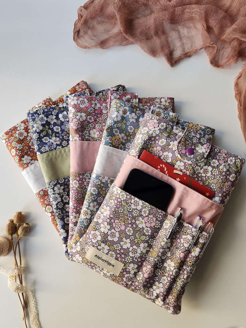May include: A collection of floral fabric pouches in various colors, including orange, navy, pink, and lavender. One pouch is open, revealing a smartphone, pens, and cards. The pouches have a button closure and a label that reads "AtelierHippie."