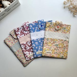 May include: Four floral fabric phone cases in various colors: pink, blue, and yellow. Each case features a different floral pattern and a button closure. The cases are handmade and have a quilted appearance. The brand name "Atelierhippie" is visible on two of the cases.