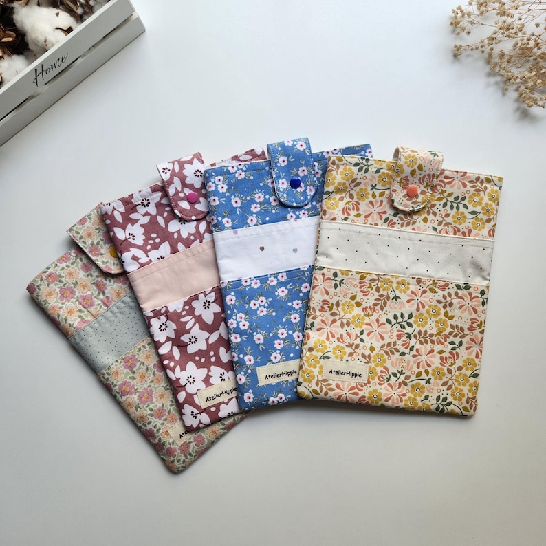 May include: Four floral fabric phone cases in various colors: pink, blue, and yellow. Each case features a different floral pattern and a button closure. The cases are handmade and have a quilted appearance. The brand name "Atelierhippie" is visible on two of the cases.