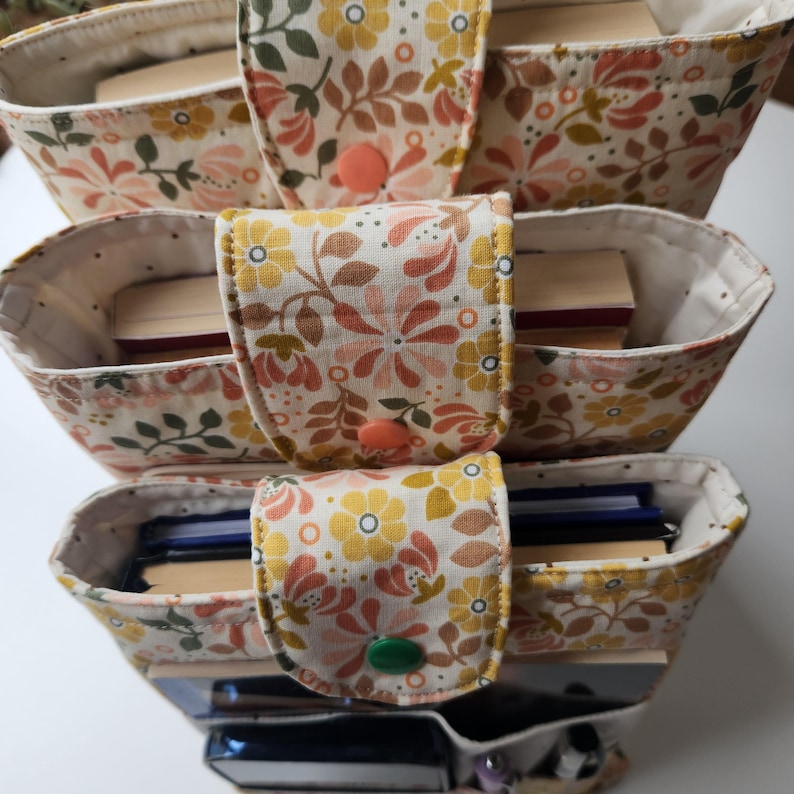 May include: A three-tiered fabric organizer with a floral pattern in cream, yellow, and peach. Each tier has a flap closure with a button. The organizer is filled with books and other small items.