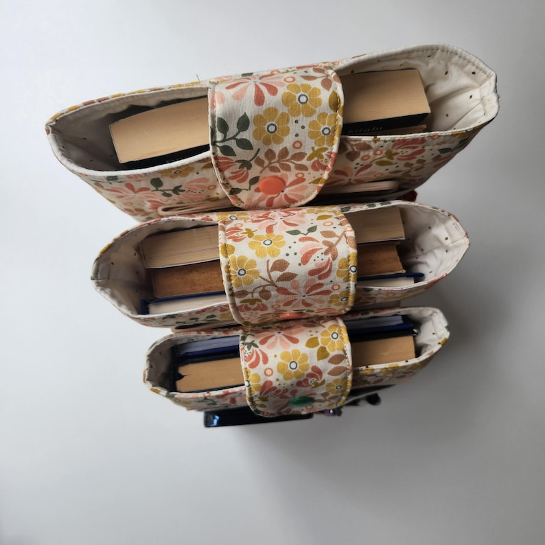 May include: Three stacked fabric book holders with a floral pattern in yellow, peach, and brown. Each holder is filled with books and secured with a matching fabric strap. The holders are on a white background.