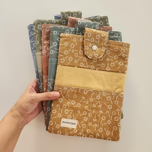May include: A stack of rectangular fabric pouches featuring a white floral pattern on various coloured backgrounds including mustard yellow, terracotta, sage green, and slate blue. Each pouch has a flap with a white snap closure and a label reading AtelierHippie.