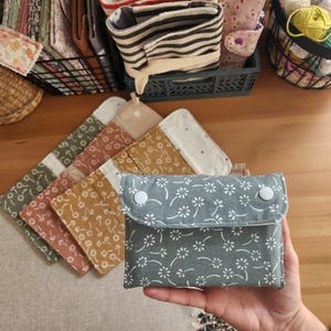 May include: A small, gray fabric pouch with a white floral pattern and two white snap closures. Several other fabric pouches in various colors and patterns are visible, along with baskets and containers.