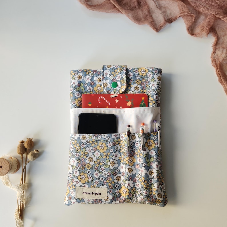May include: A floral print fabric phone case with a button closure. The case is blue with yellow and white flowers. Inside, there is a black smartphone, a red card with a Christmas design, and pens. The label reads "AtelierHippie".