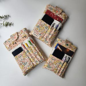 May include: Three floral fabric organizers with button closures. Each organizer has pockets for pens, phones, and cards. The fabric features a pattern of yellow flowers, green leaves, and red accents on a light pink background. The text "Atelierhippie" is visible on one.