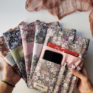 May include: A collection of floral print fabric cases in various colors, including pink, blue, and brown. One case is open, revealing a phone, pens, and a card. The cases have a button closure and a label that reads "AtelierHippie."
