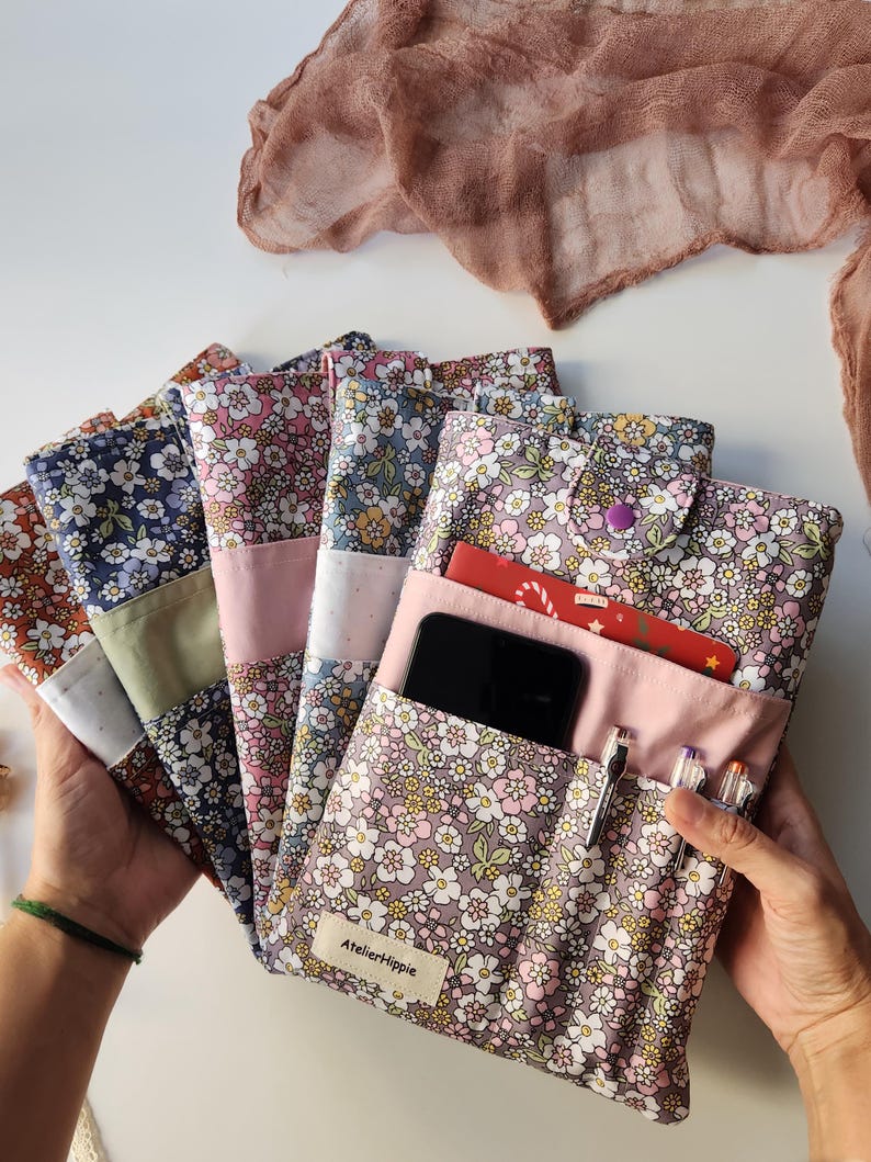 May include: A collection of floral print fabric cases in various colors, including pink, blue, and brown. One case is open, revealing a phone, pens, and a card. The cases have a button closure and a label that reads "AtelierHippie."
