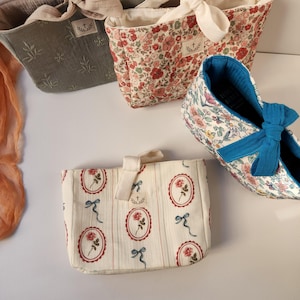 May include: An assortment of fabric tote bags and a pair of shoes. The bags feature floral and botanical patterns in shades of red, pink, and green. One bag is a solid gray-green color. The shoes have a blue bow.