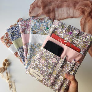 May include: A collection of floral fabric pouches in various colors, including orange, blue, and pink. One pouch is open, revealing a smartphone, pens, and a red card. The pouches have a button closure and a label that reads "Atelier Hippie."