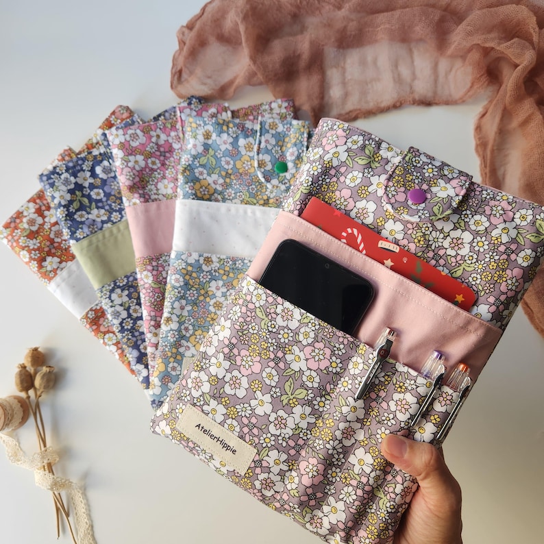 May include: A collection of floral fabric pouches in various colors, including orange, blue, and pink. One pouch is open, revealing a smartphone, pens, and a red card. The pouches have a button closure and a label that reads "Atelier Hippie."