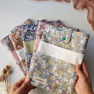 May include: A collection of floral patterned fabric cases in various colors, including blue, orange, and pink. Each case features a white accent and a button closure. The cases are arranged in a fan shape, with the text "Atelier Hippie" visible on one.