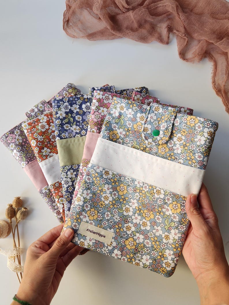 May include: A collection of floral patterned fabric cases in various colors, including blue, orange, and pink. Each case features a white accent and a button closure. The cases are arranged in a fan shape, with the text "Atelier Hippie" visible on one.