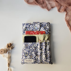 May include: A navy blue floral print organizer with a light blue button closure. The organizer holds a smartphone, pens, and a red card. The text "AtelierHippie" is on a small white label. The background is white with a pink scarf.