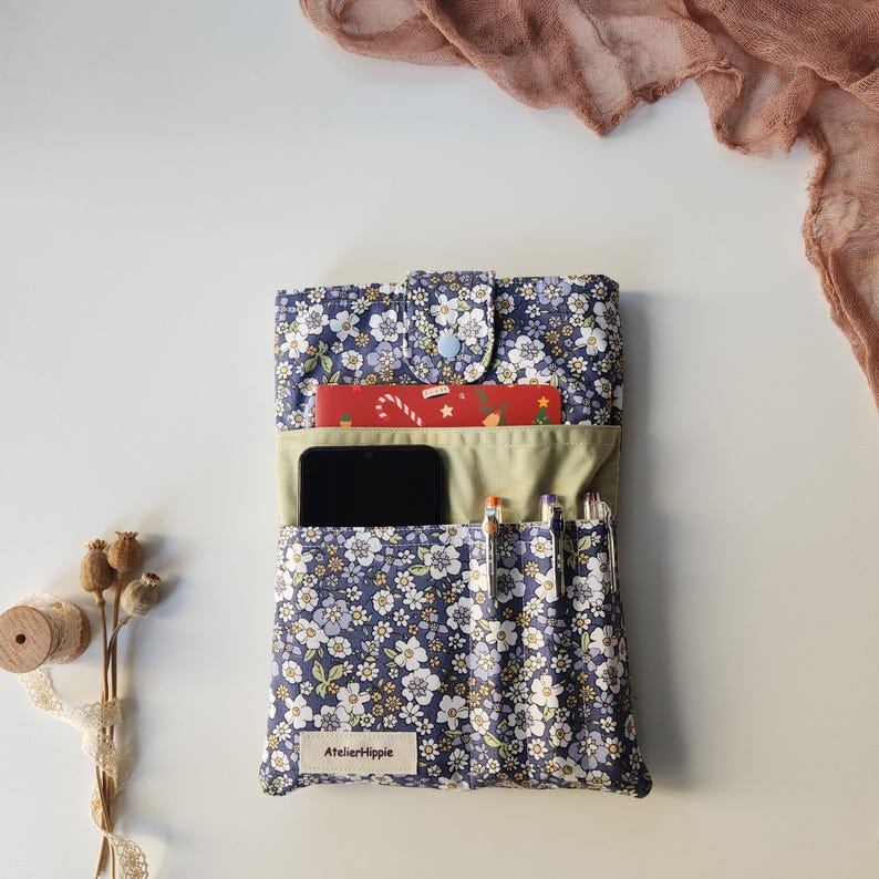 May include: A navy blue floral print organizer with a light blue button closure. The organizer holds a smartphone, pens, and a red card. The text "AtelierHippie" is on a small white label. The background is white with a pink scarf.