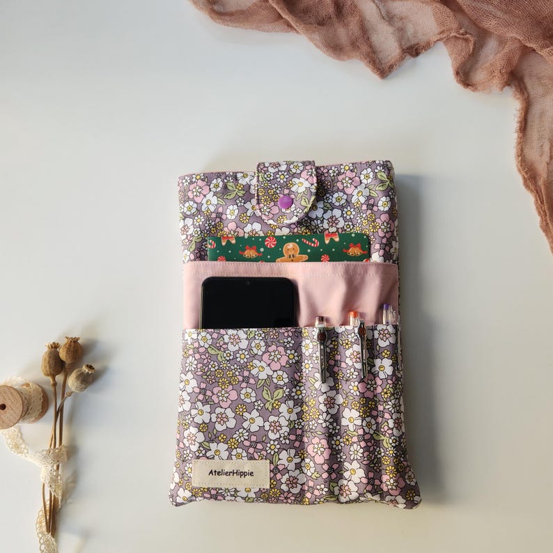May include: A floral print tablet case with a button closure. The case is lavender with a pattern of small white and pink flowers. It has pockets for a phone, pens, and a small card. The text "AtelierHippie" is visible on the bottom.