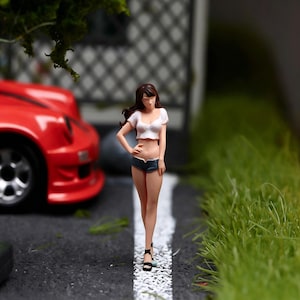 May include: Miniature figurine of a woman in a white crop top, denim shorts, and black heels, standing on a road with a white stripe. A red sports car is in the background, along with green grass and a building.